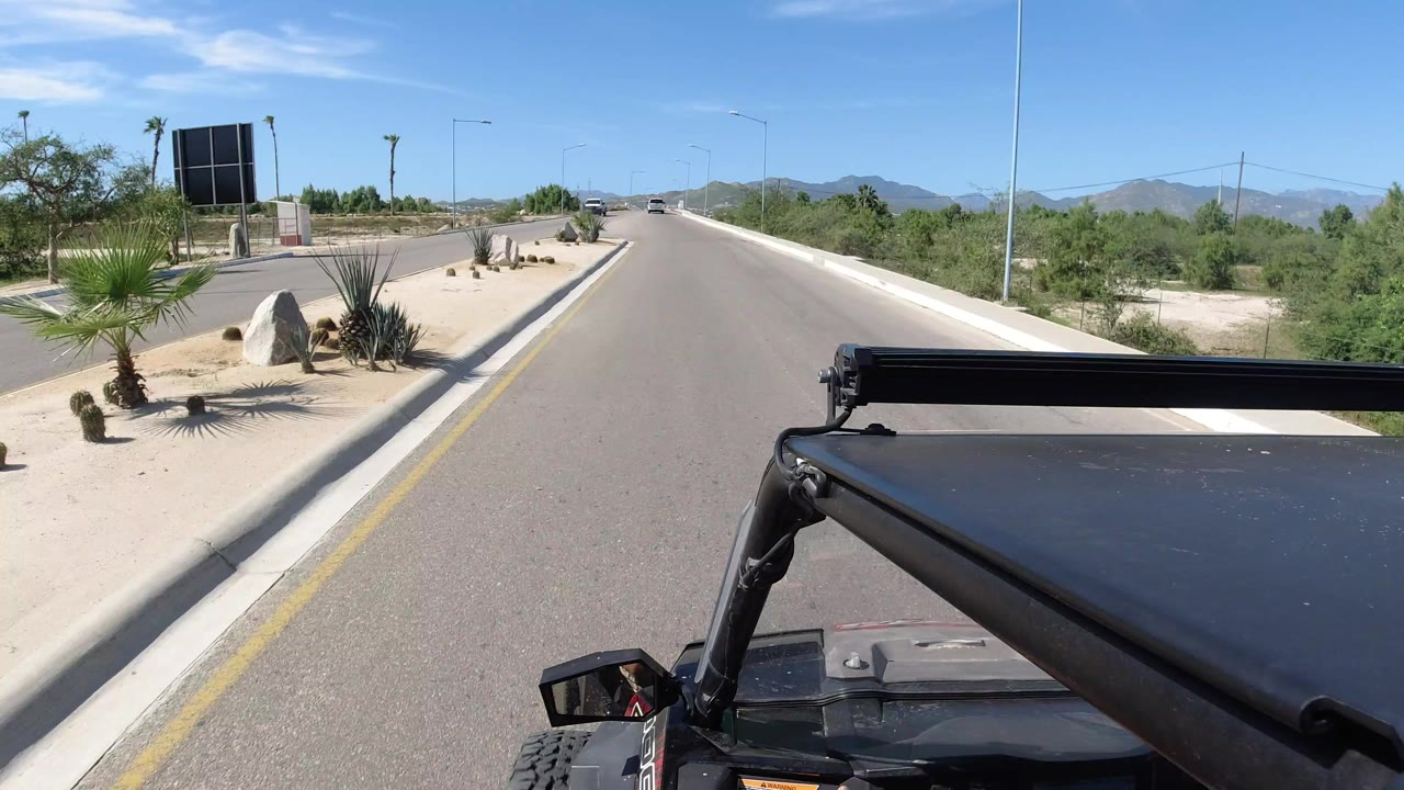 A Los Cabos road view from a Wrangler shows the black roof and mirror in the foreground, a paved two-way road ahead, desert plants, palms, distant hills, and no visible people.