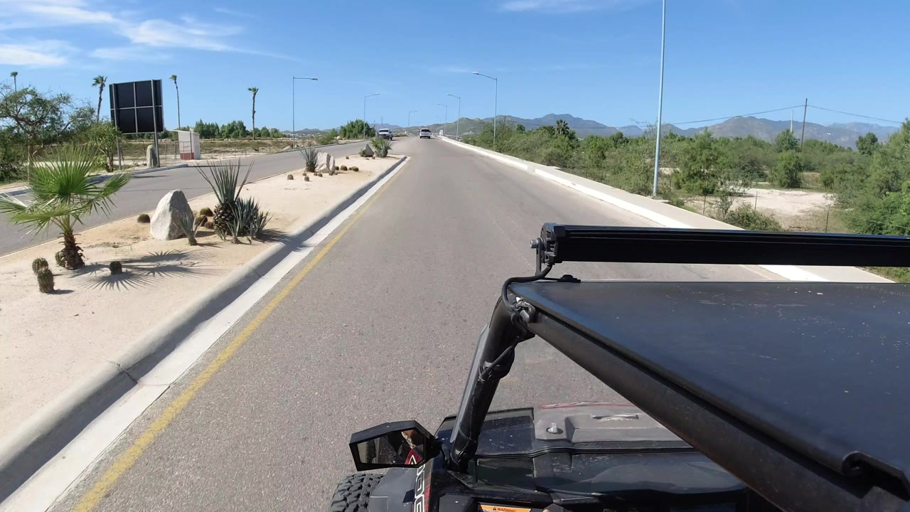 A forward view over the Wrangler roof shows the Los Cabos two-way road continuing past roadside rocks, palms, desert shrubs, streetlights, distant vehicles, and no visible people.