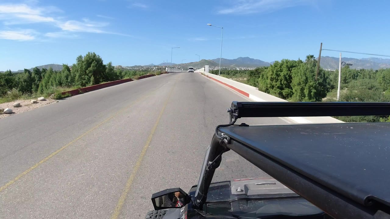 Jeep-mounted view of a paved road in Los Cabos, Mexico, with black vehicle hardware in the foreground, green trees at the roadside, distant mountains, and no visible people.