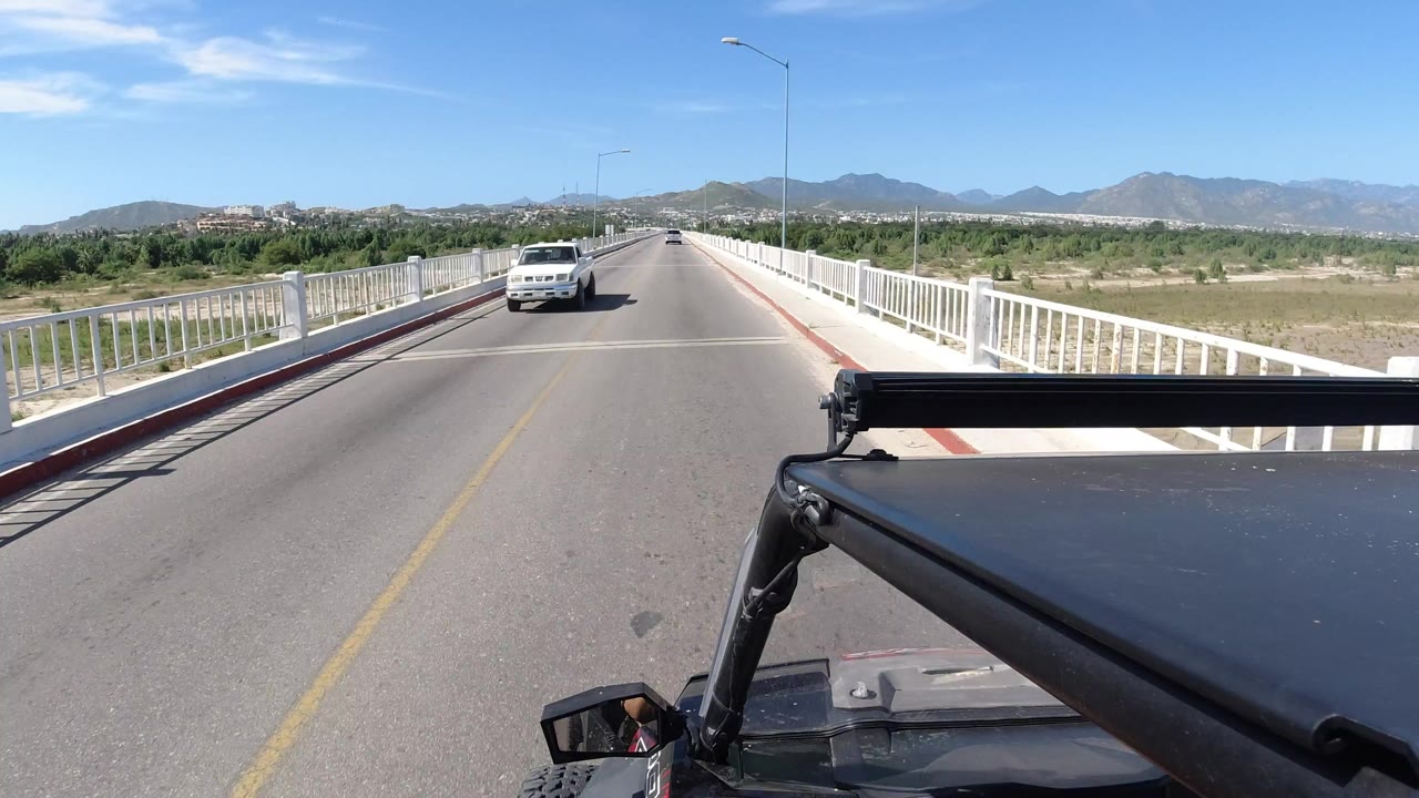 Start of the Los Cabos, Mexico bridge drive, with an opposite-direction Jeep approaching on the left side of the road, white railings on both sides, distant mountains, and no visible people.
