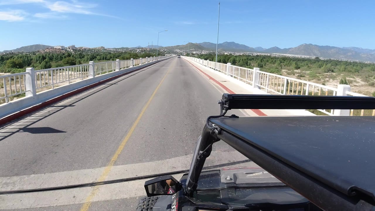 One second into the Los Cabos, Mexico drive, the road remains centered between white railings, with green scrubland and a low mountain range under bright sun, and no clearly visible people.