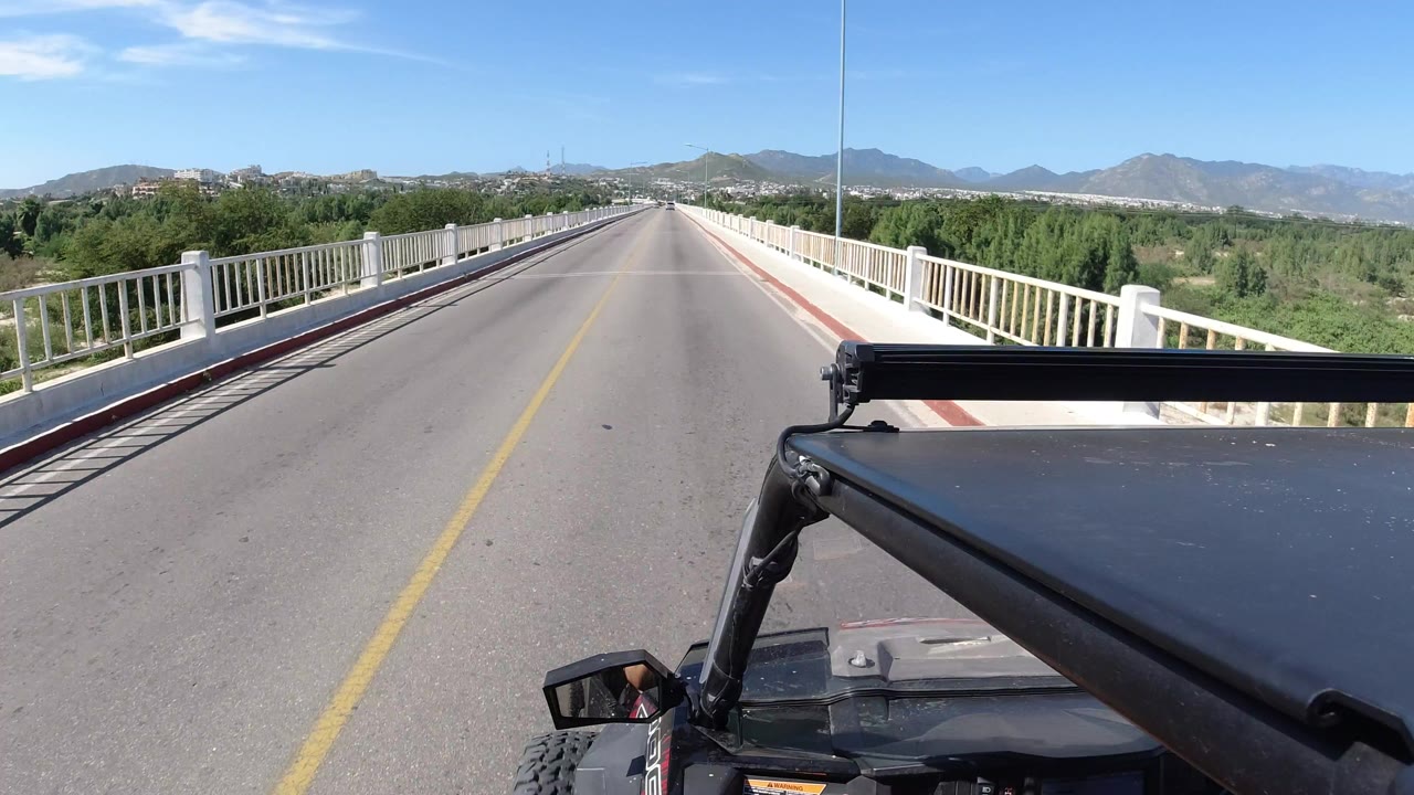 Jeep hood on a sunny bridge road with white railings and mountains ahead.