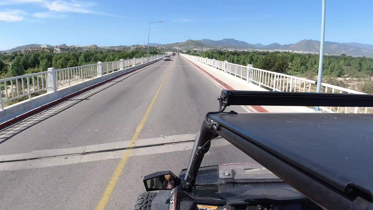 Vehicle-mounted view down a sunny bridge road with white railings and mountains ahead.