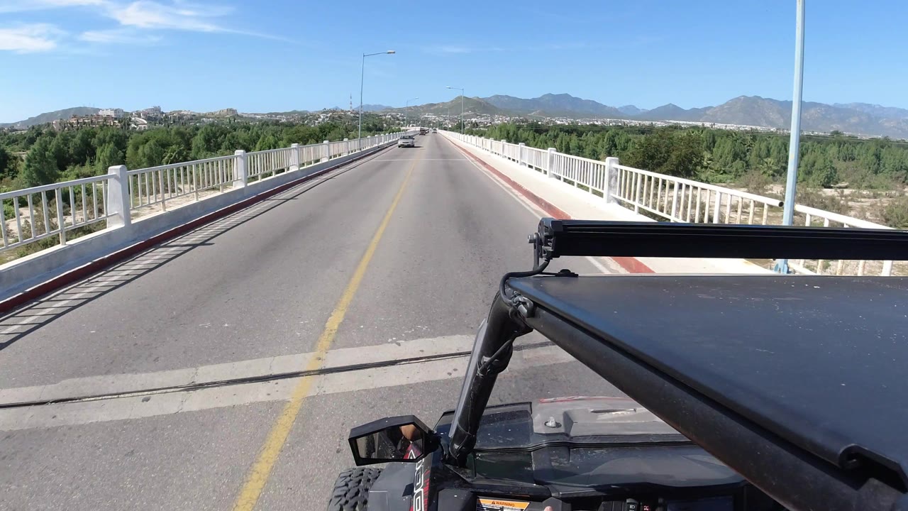 At 0:01, the Los Cabos bridge road remains centered ahead, with the filming vehicle hardware low in frame, white railings on both sides, distant traffic, green vegetation, and no clearly visible people.