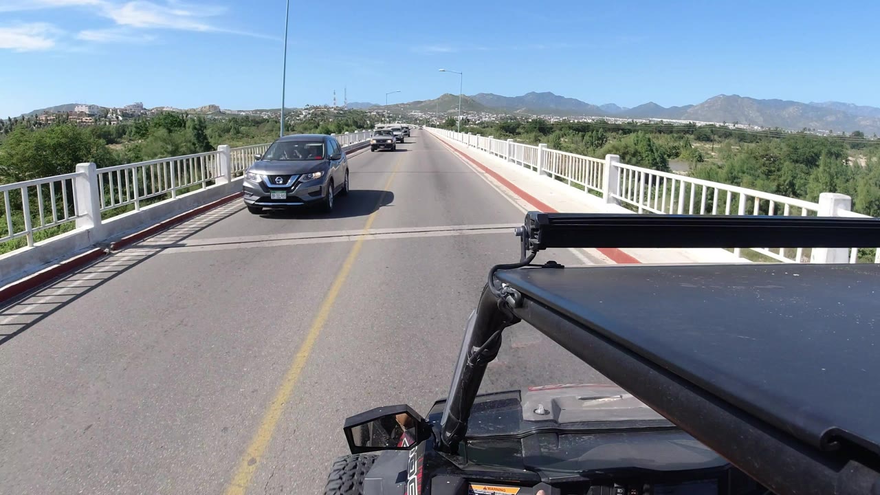 A dark car draws closer on the Los Cabos bridge while the foreground vehicle roof and mirror remain visible against white railings, trees, hills, and no visible people.