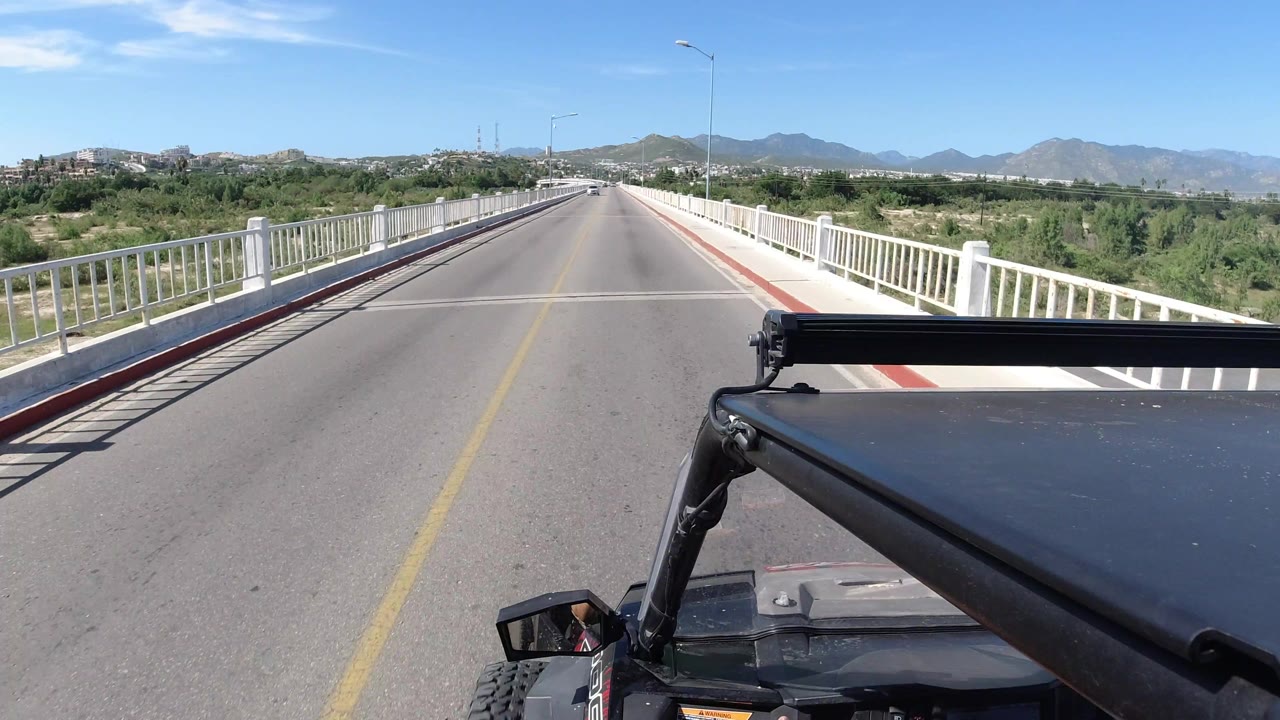 The Wrangler remains low in the foreground as the Los Cabos bridge road runs straight toward low hills and distant mountains, with no visible people.