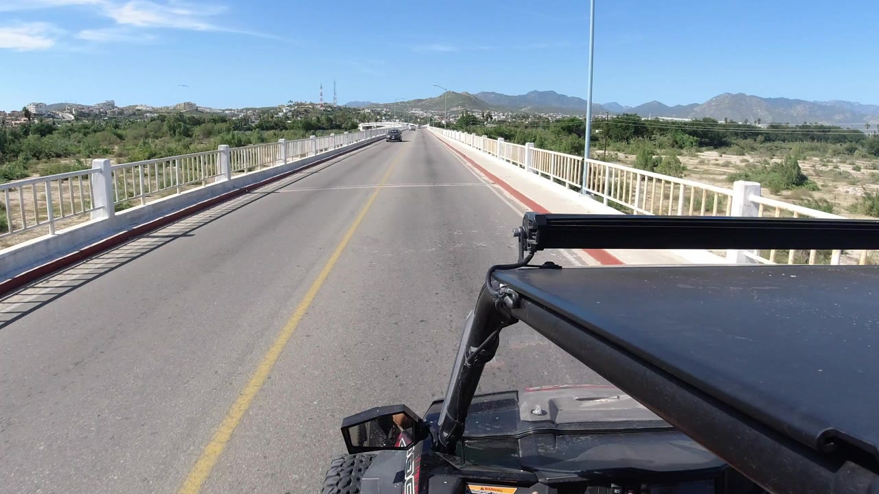 Opening frame of a vehicle-mounted road view on a bridge in Los Cabos, Mexico, with white railings, dry terrain, distant mountains, blue sky, and no visible people.