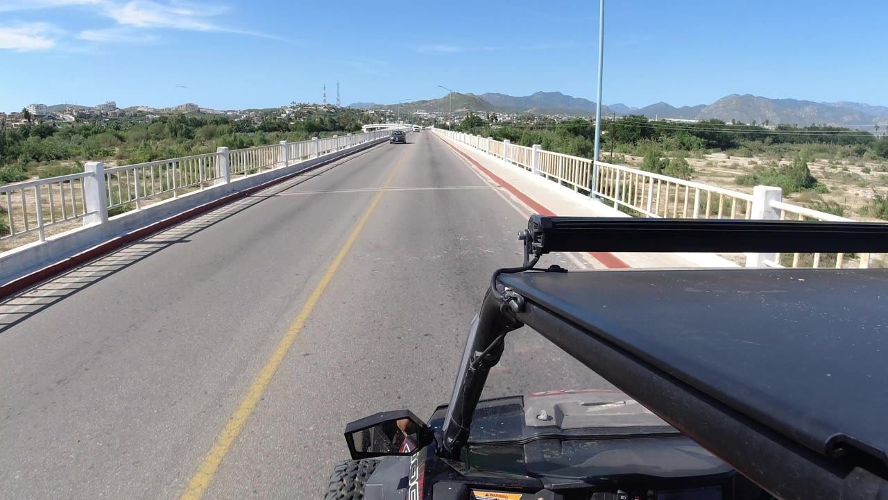 Forward road view from a mounted camera in Los Cabos, Mexico, showing the bridge rails converging ahead, a vehicle in the distance, scrubland below, and no visible people.