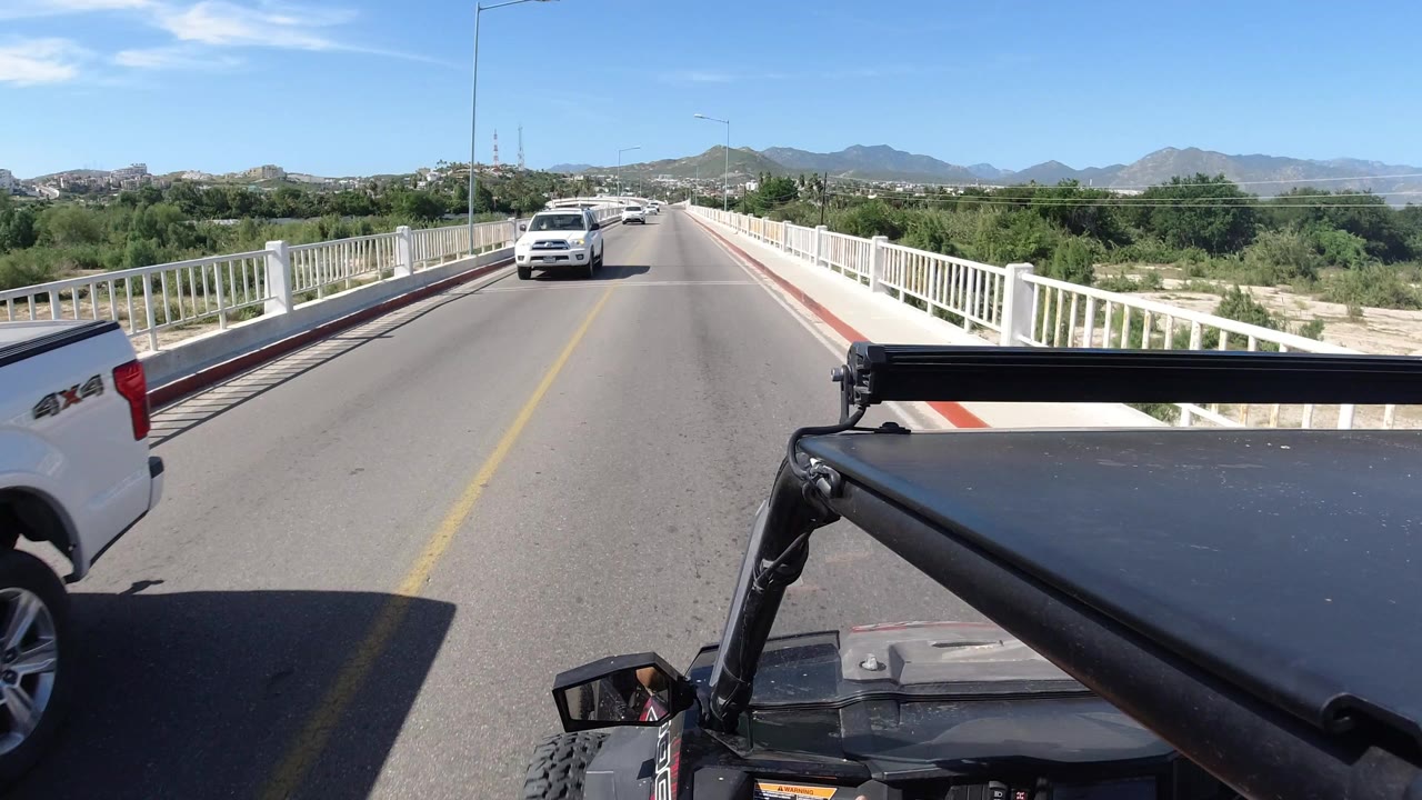 Los Cabos bridge road seen from a vehicle, with a white SUV ahead in the opposite lane, railings on both sides, roadside greenery, low mountains, and no visible people.