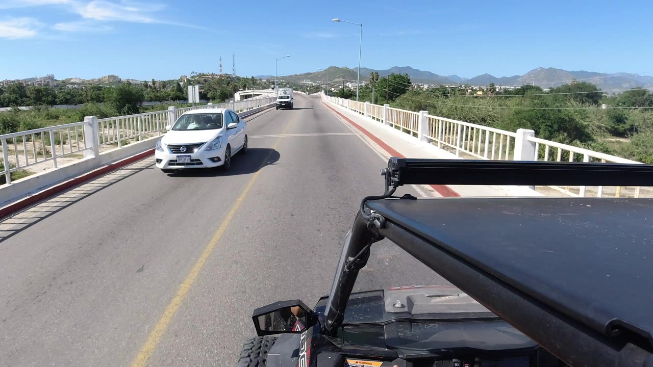 The white car is closer in the lane on a Los Cabos bridge, with the foreground vehicle hardware at lower right, roadside trees, mountain ridges, and no visible people.