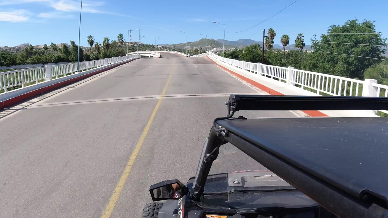 Forward view from a vehicle on a Los Cabos road, with the bridge railings converging toward distant hills and no visible people.