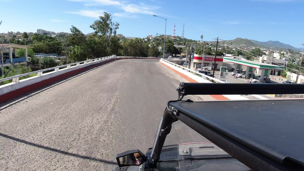 Opening Los Cabos frame from a Jeep-mounted camera, showing the vehicle roof hardware, a quiet paved road, white guardrails, trees, hillside buildings, distant hills, and no visible people.