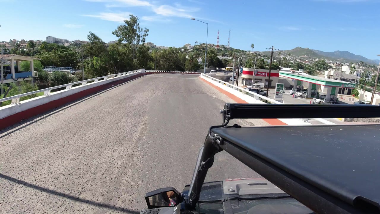 Los Cabos road view with Jeep roof hardware in the foreground, a curving street ahead, roadside trees, utility lines, hillside structures, a Circle K storefront at right, and no visible people.