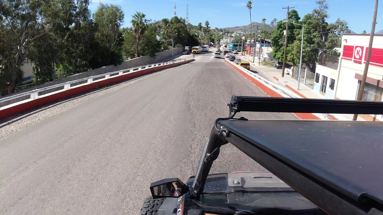 The Jeep foreground remains steady while the Los Cabos road leads downhill between trees, red-and-white curbs, parked vehicles, and low commercial buildings, with no visible people.