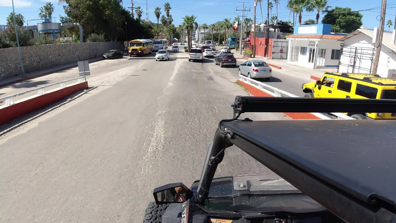 Forward-facing view from a Wrangler on a sunlit Los Cabos road, with cars ahead, a yellow bus on the left, palm trees, roadside walls, and no visible people.