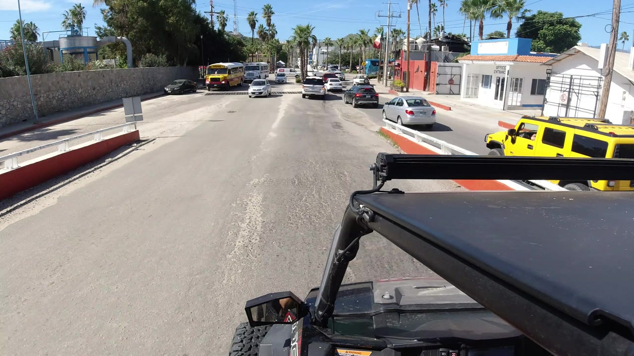 The Wrangler roof and roll bar frame a Los Cabos street view with cars moving ahead, a yellow bus farther left, a white roadside building, palm trees, and no visible people.