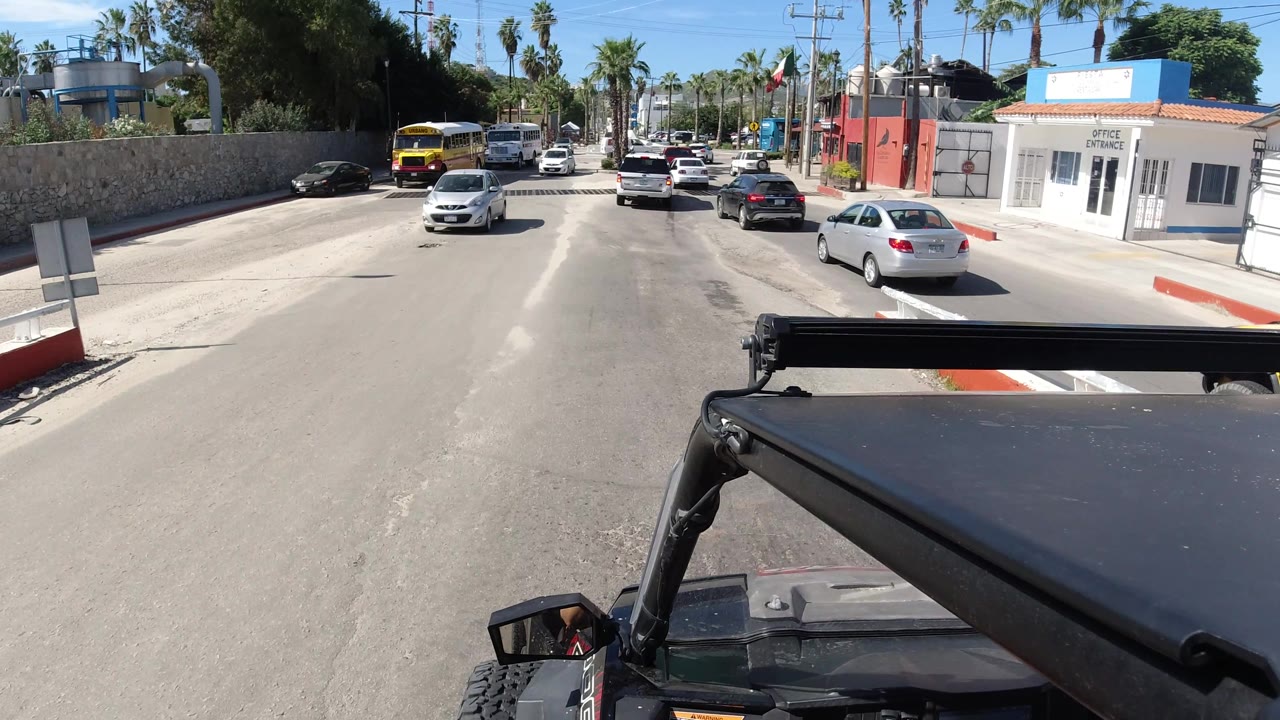Road traffic passes a white office entrance building with palms and the Wrangler roof in foreground.