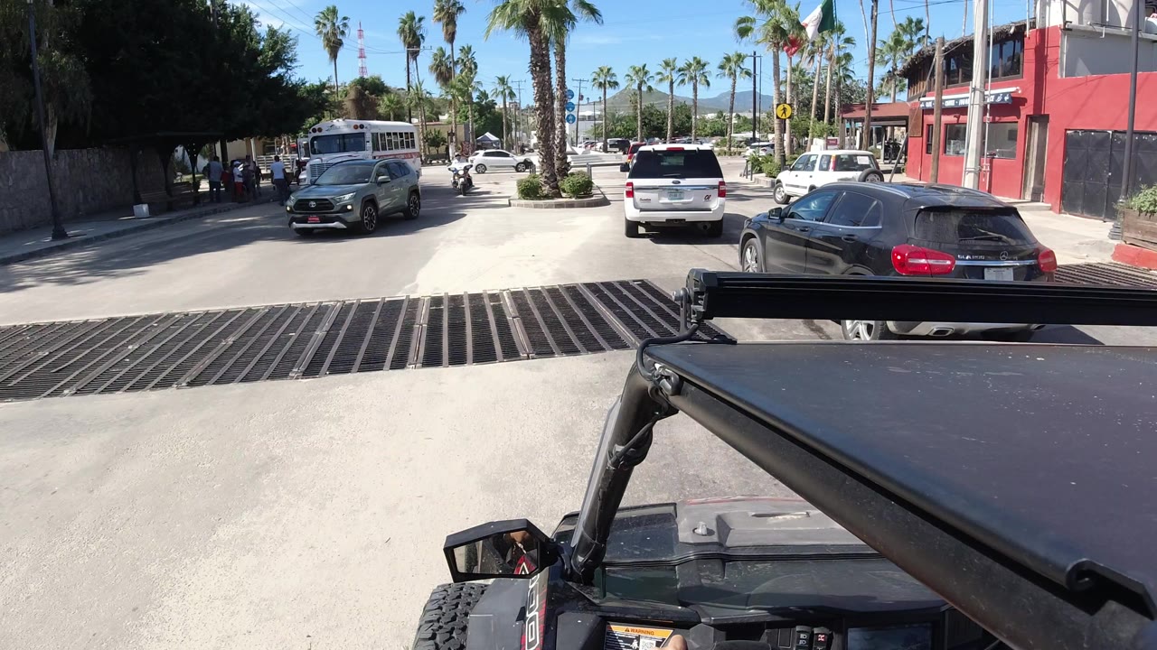 Vehicle viewpoint facing a sunny street with cars, a bus, palms, and a red building.