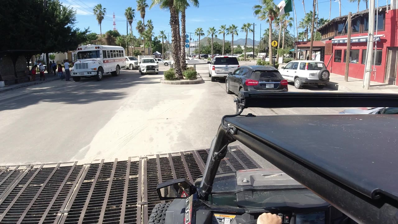 Driver-level view in Los Cabos, Mexico with black vehicle hardware in the foreground, cars ahead, a white bus to the left, palm trees, and a red storefront at the right.