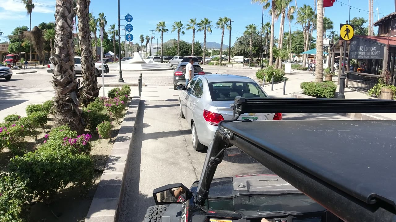 Start of a Los Cabos driving view with a vehicle roof in the foreground, a parked silver sedan ahead, palm trees, bollards, and bright street landscaping, with no clearly visible people.