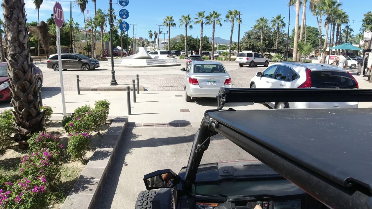 Jeep roof and frame in the lower foreground face a sunlit Los Cabos street with parked cars, palm trees, bollards, a stop sign, and distant plaza structures.