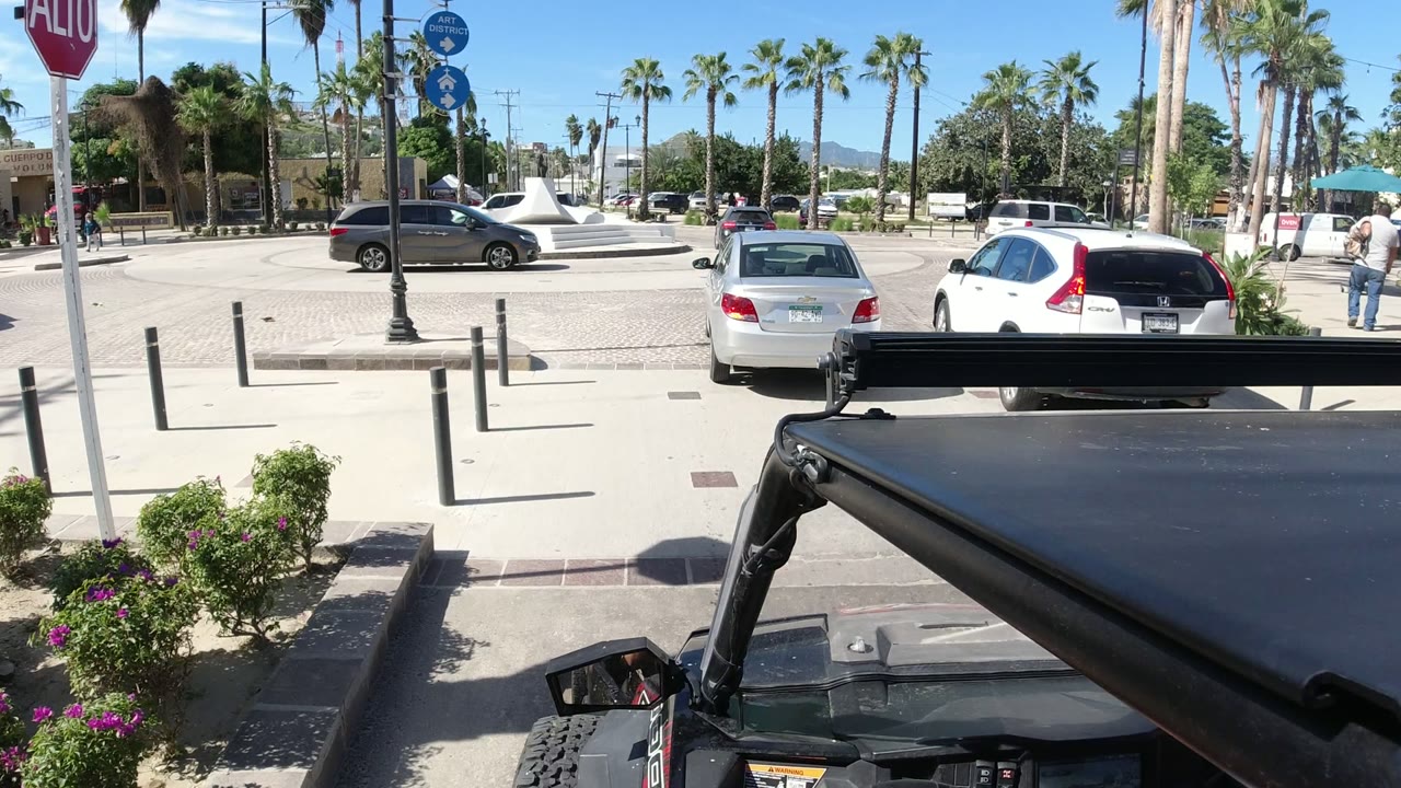 The Jeep foreground remains at the bottom of the frame as a black car, white vehicle, palm trees, bollards, and a white sculptural plaza feature appear across the road in Los Cabos.