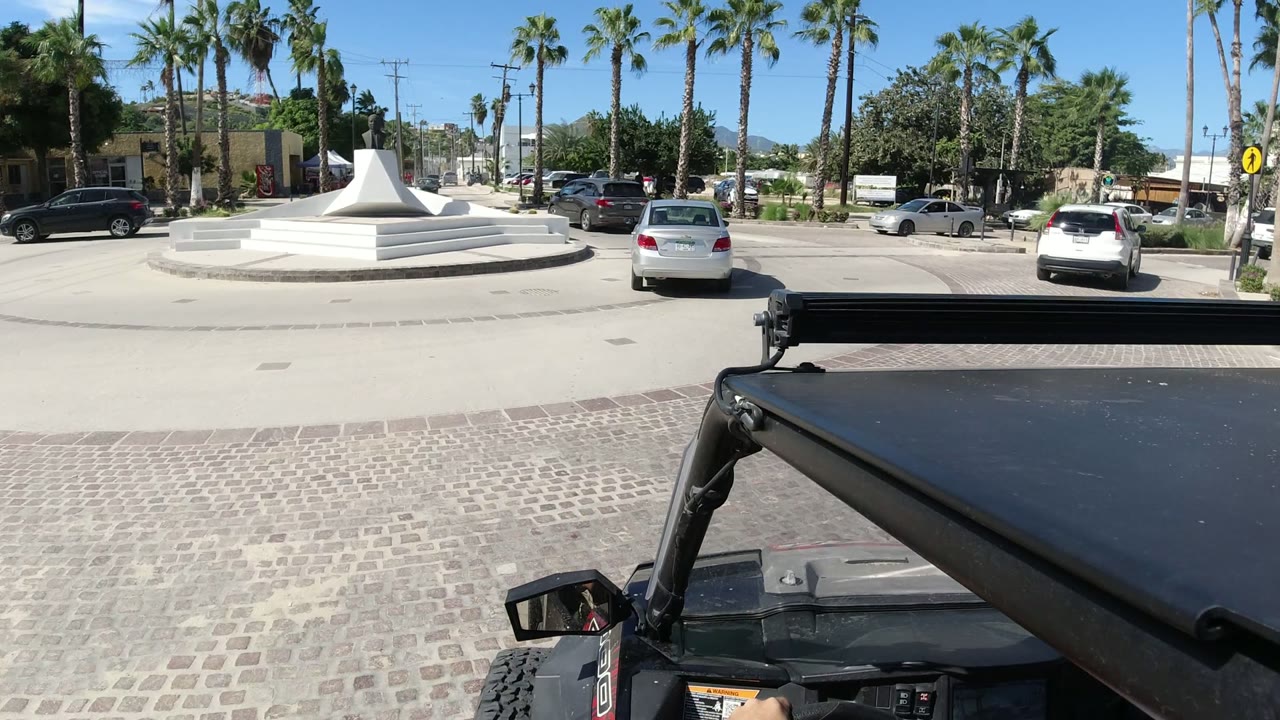 Street-level view in Los Cabos, Mexico with a dark vehicle roof in the foreground, a white stepped roundabout monument to the left, cars on the road, palm trees, and no visible people.
