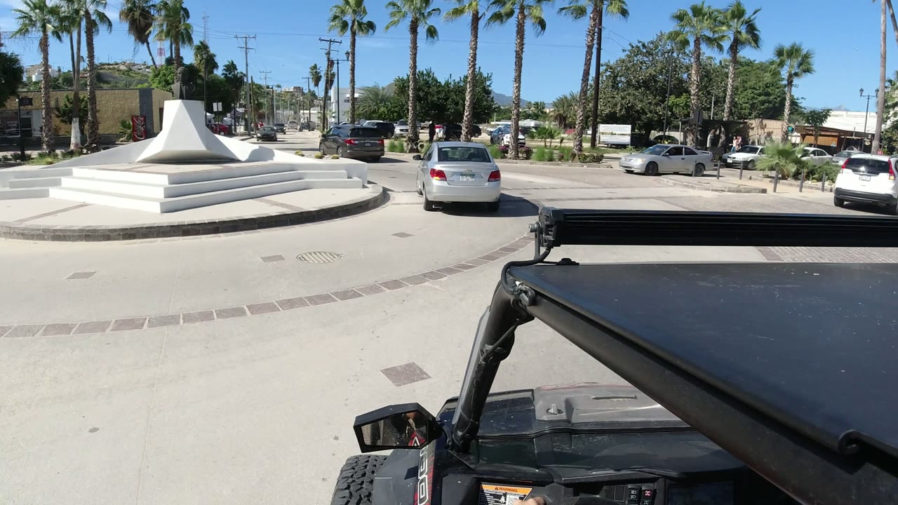 Los Cabos roundabout view with the white monument more centered, the vehicle roof cutting across the lower frame, parked and moving cars beyond, palm trees, and no visible people.
