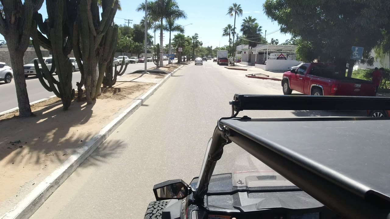 Los Cabos street POV from a Wrangler, passing a red pickup, white parked cars, palms, and low buildings in strong sunlight.