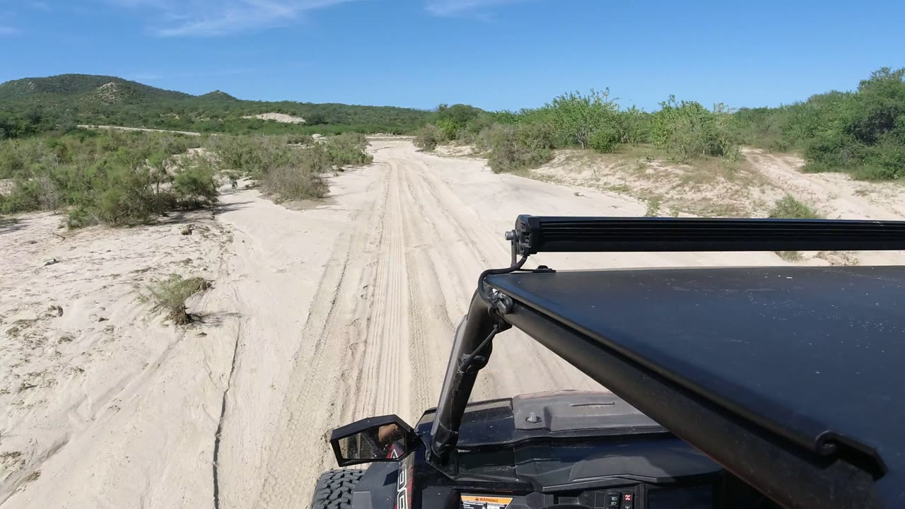 Black off-road vehicle hood beside a sandy desert road with shrubs and low mountains.