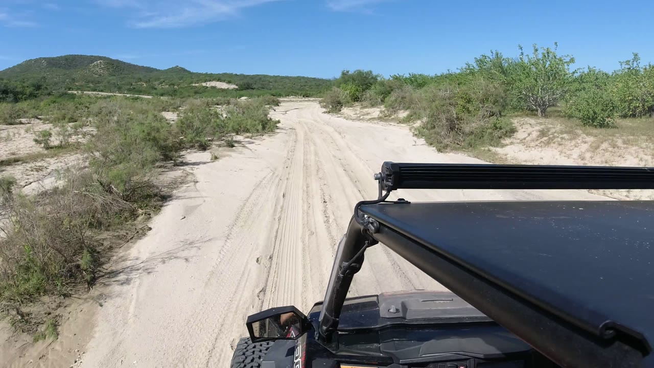 One second in, the Los Cabos sandy track runs straight ahead between low green brush, while the black vehicle hood and light bar occupy the lower right foreground and no full person is visible outside the vehicle.