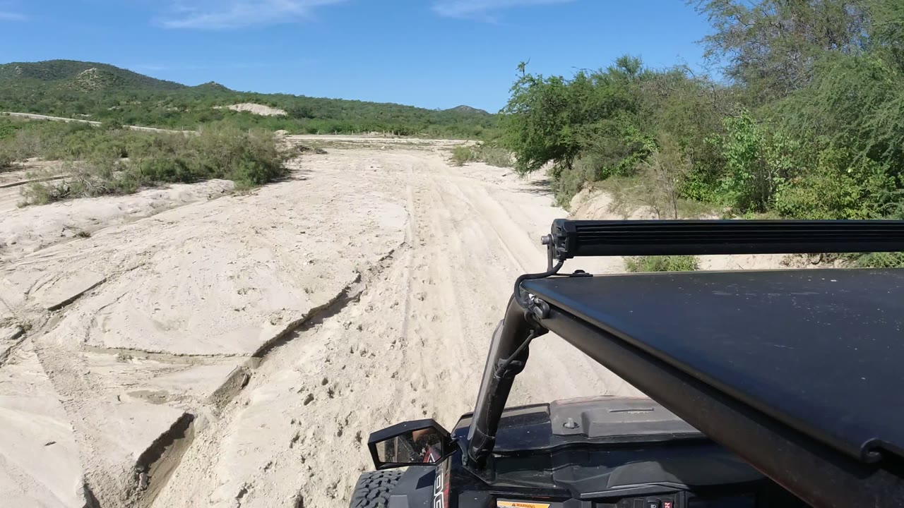 Opening frame in Los Cabos, Mexico, looking past a black off-road vehicle hood toward a sandy wash, green shrubs, low hills, blue sky, and no recognizable people.