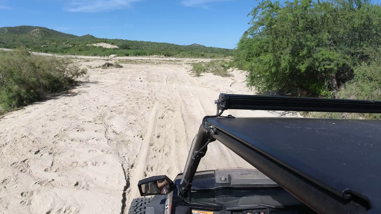 Second frame in Los Cabos, Mexico, with the black vehicle hood and roof bar low in frame, a pale sandy track ahead, scrub vegetation at right, distant hills, and no recognizable people.