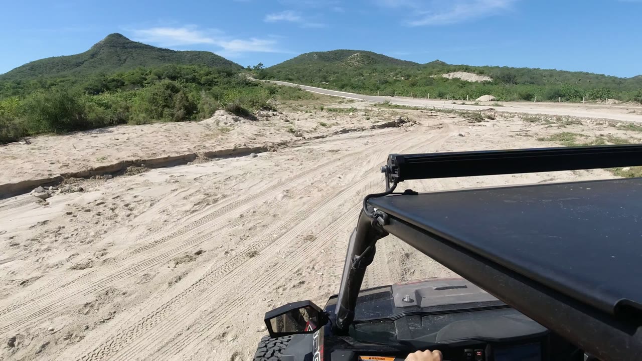 The Jeep approaches a sandy wash in Los Cabos, Mexico, with a pale roadside crossing and low green hills beyond, no visible people.