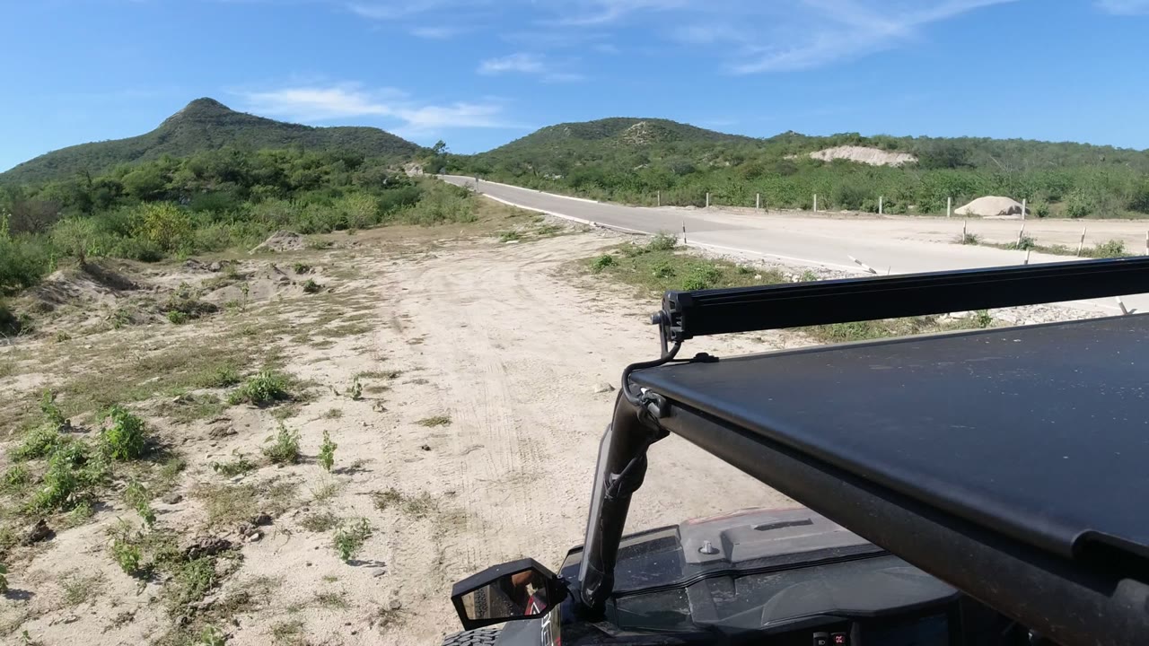 Second frame shows the black vehicle roof in the foreground with a broad sandy shoulder, paved road, roadside posts, brush, and hills in Los Cabos, Mexico.