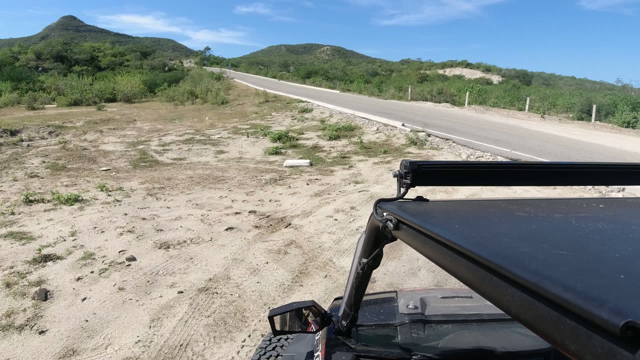 Off-road vehicle foreground beside a paved desert road and low green hills.