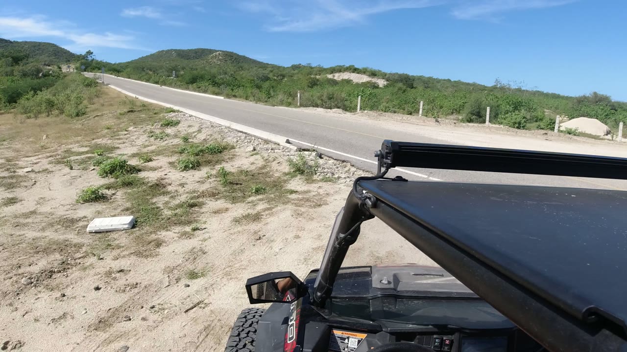 A black off-road vehicle roof and mirror sit in the foreground as a paved Los Cabos road runs past sandy ground, shrubs, roadside posts, and low hills, with no visible people.