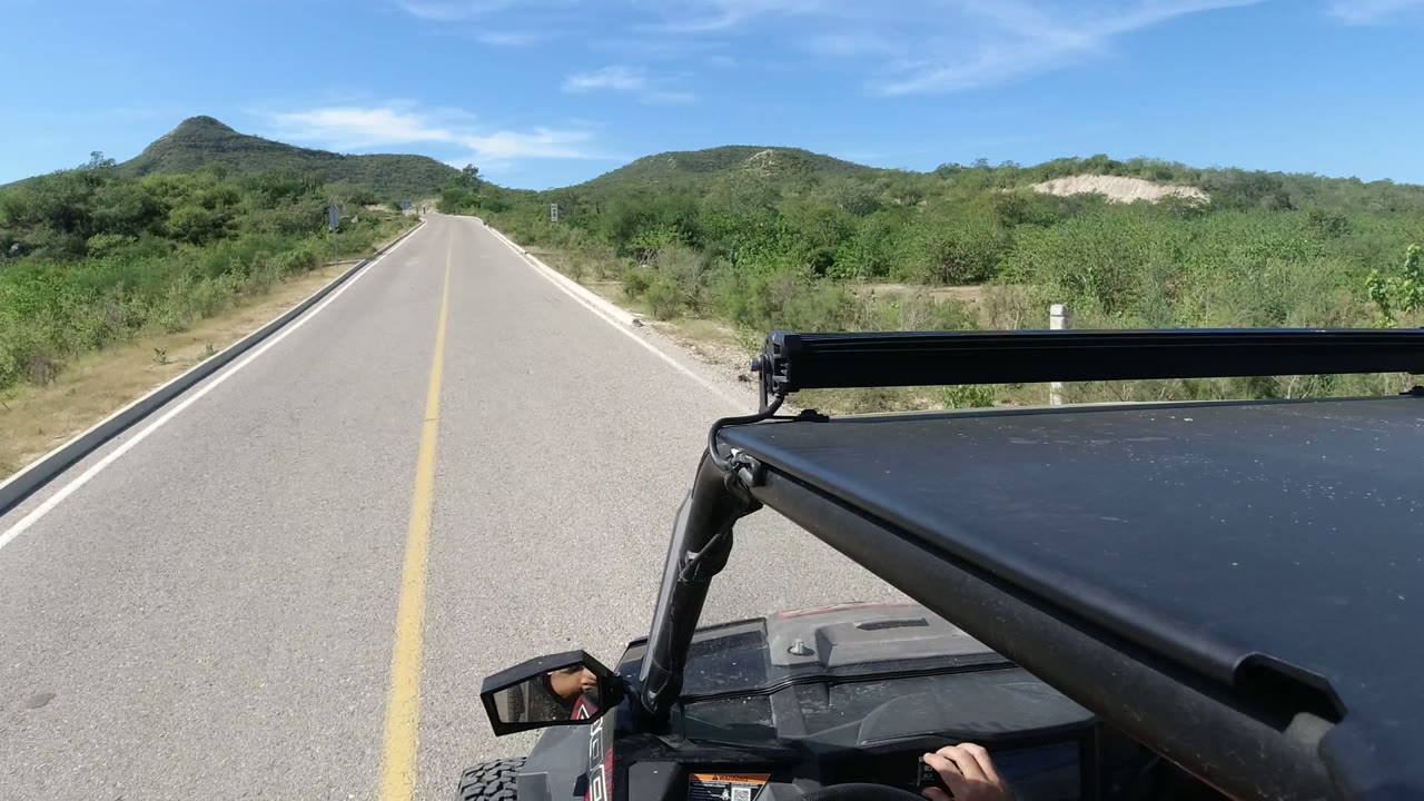 The Los Cabos road remains centered as the Wrangler foreground shifts slightly lower right, with scrub vegetation and rounded hills under a clear blue sky and no recognizable people.