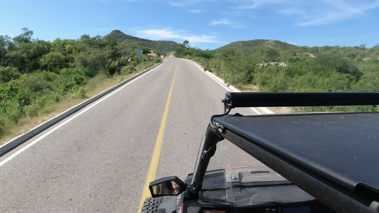 Two-lane road through green roadside brush with vehicle roof in foreground