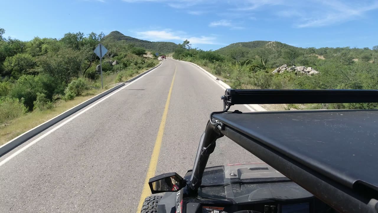 Forward-facing Los Cabos, Mexico driving frame showing a roadside sign on the left shoulder, dense green vegetation, distant hills, and no visible people.