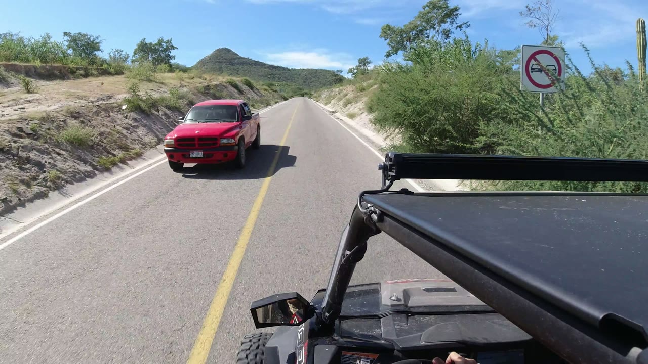 The red vehicle passes near the camera on the Los Cabos road, with the Jeep foreground hardware at lower right, pale roadside banks, green shrubs, and no visible people.