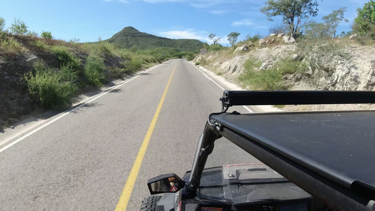 Forward road view with vehicle frame, green roadside brush, and a hill ahead.