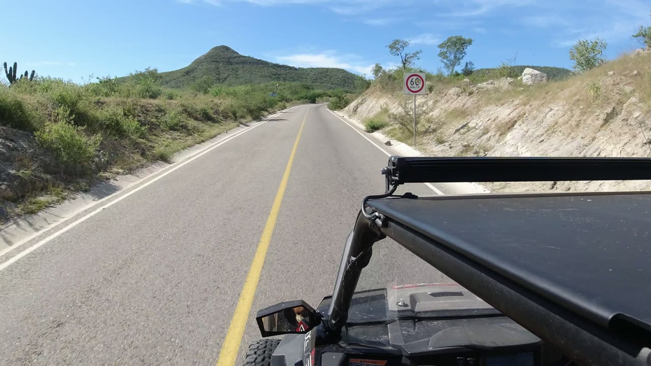Los Cabos road view with the vehicle frame at lower right, a circular roadside speed sign on the sandy shoulder, scrub vegetation, a rounded hill ahead, blue sky, and no visible people.