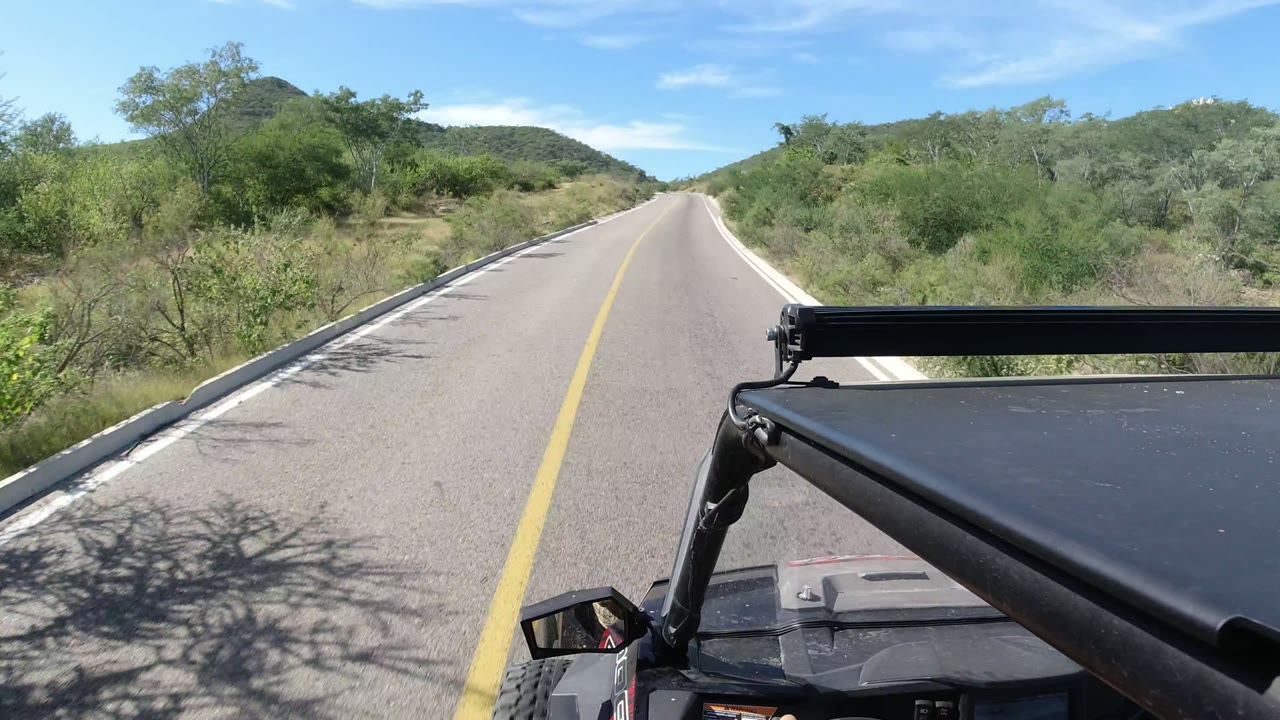 Opening frame from Los Cabos, Mexico shows a paved road ahead with scrubby hills, a yellow center line, the RZR roof edge in the foreground, and no visible people.