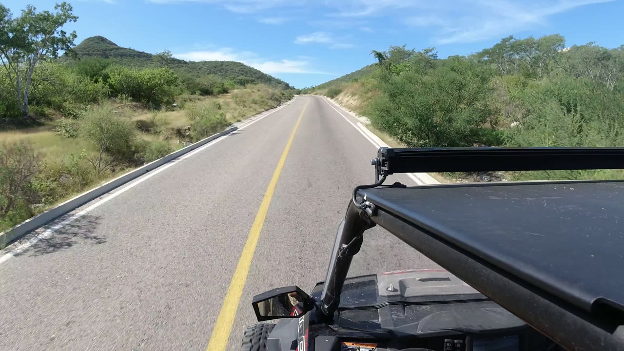 At 0:01, the Los Cabos road continues between green roadside brush and low hills, with the RZR frame crossing the lower right foreground and no visible people.