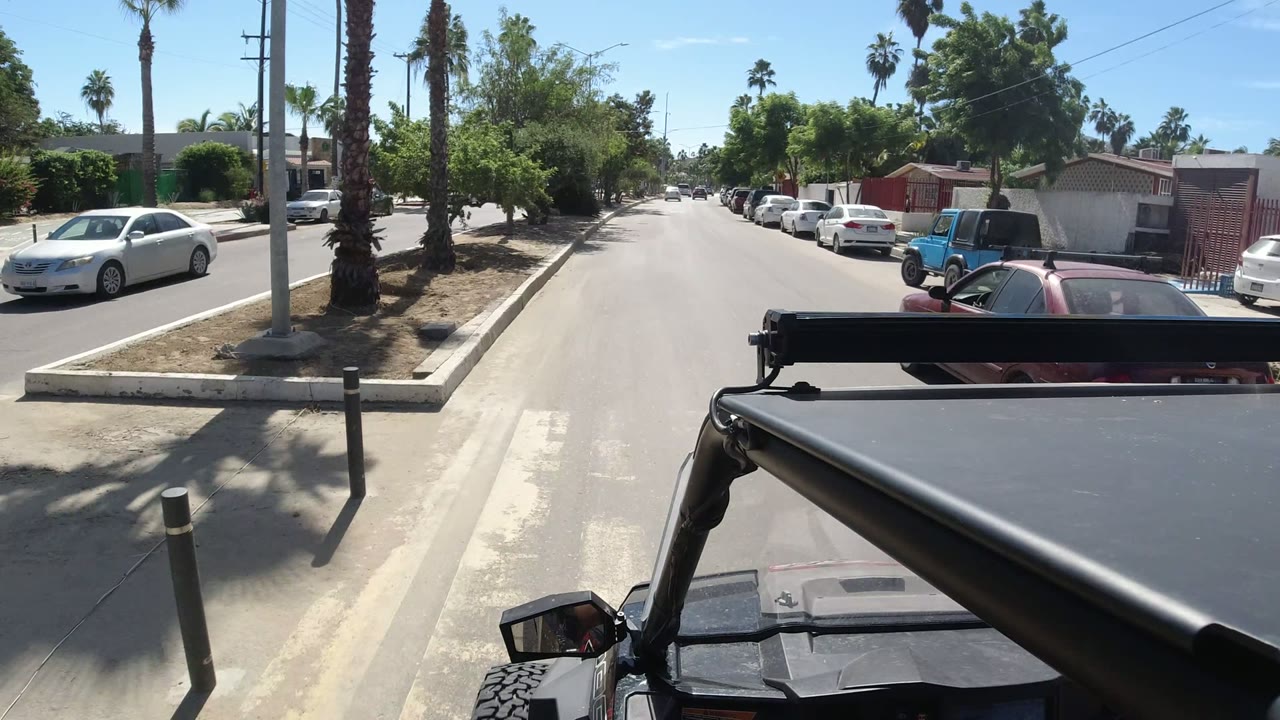 The Los Cabos street continues ahead from the Wrangler, with a white car on the right, palms and trees overhead, low residential walls, and no clearly visible people.