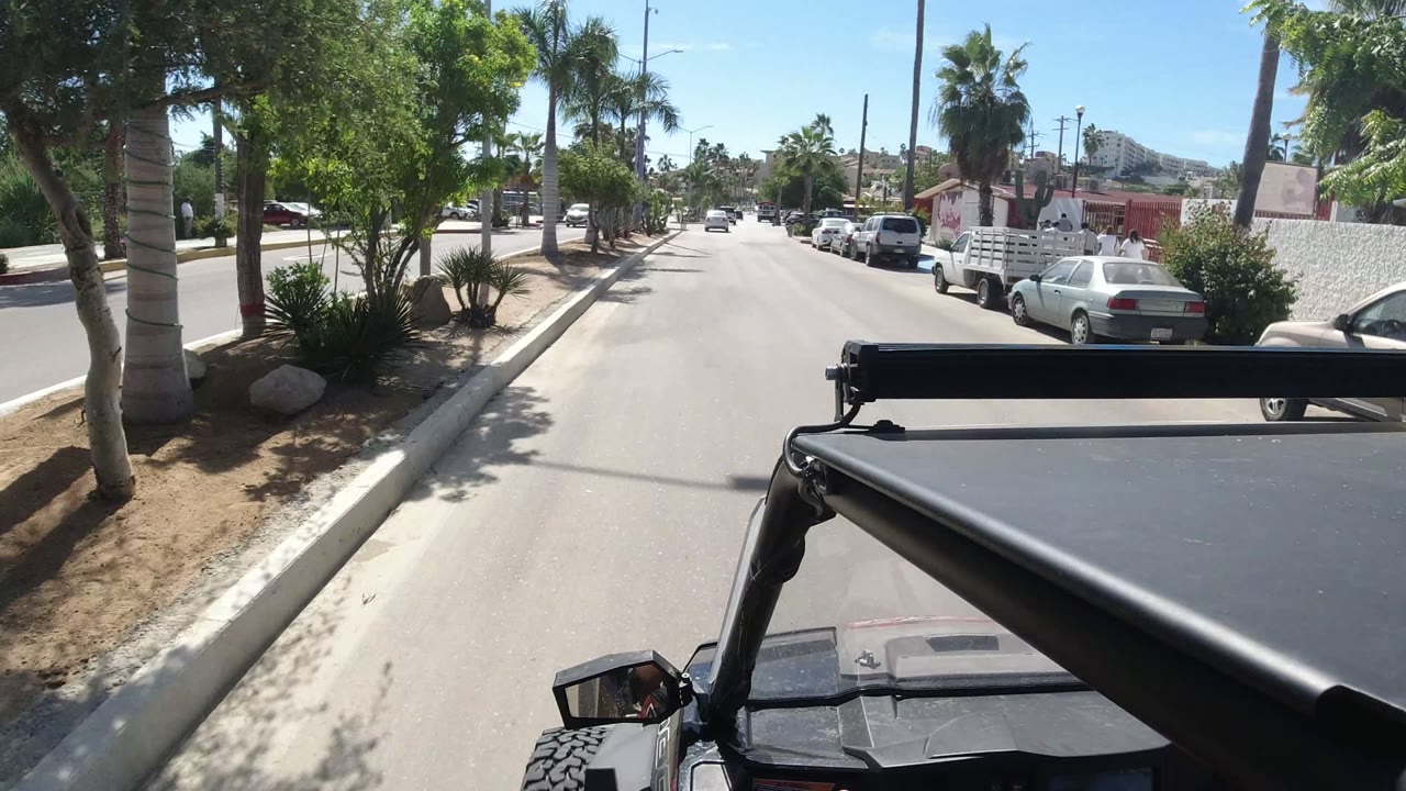 Forward roadway view with palms, parked cars, and a low wall on the right.