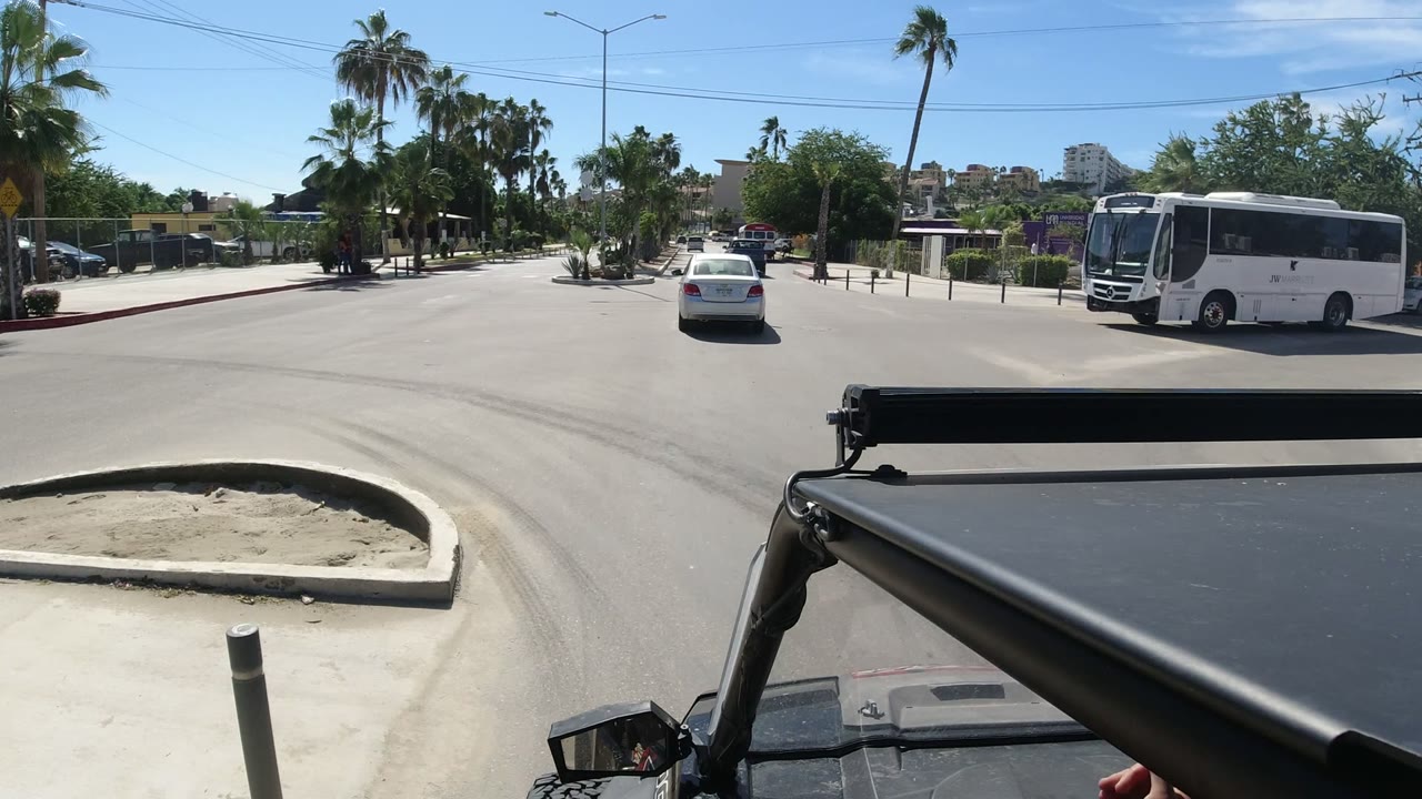 Los Cabos, Mexico crossroad seen from a Wrangler, with the white bus closer on the right, a small white car ahead, palms, road islands, and no visible faces.