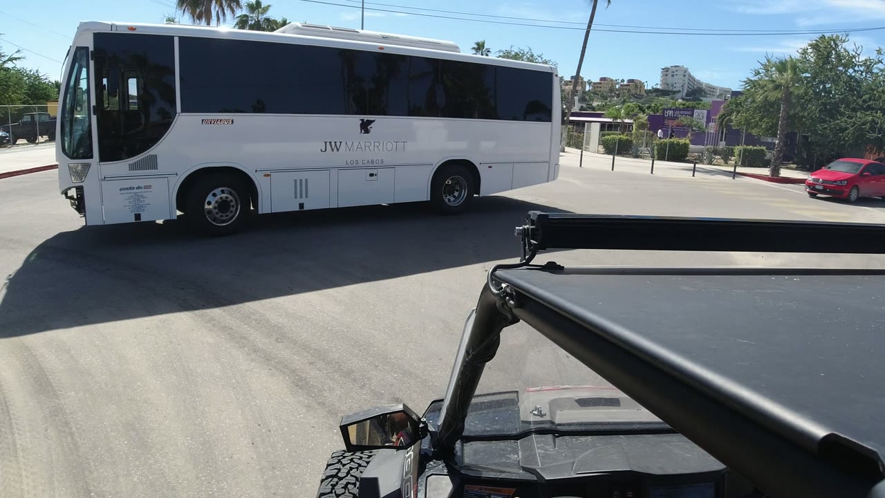 One second in, the white Marriott Los Cabos shuttle bus fills the center of the Los Cabos street view, with the open vehicle roof line in the foreground and palms across the road.