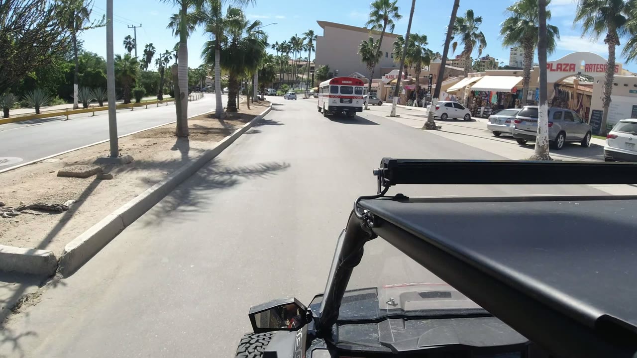 Los Cabos street view one second in, with the camera framed by a dark vehicle roof edge, a bus ahead, shop signs on the right, parked cars, and no clearly visible people.